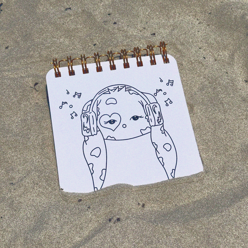 A small spiral-bound notepad lying on sand, featuring a black line drawing of a face with headphones and small music notes on a white sheet of paper.